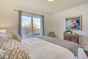 A bedroom with a large bed, a window, a painting on the wall, and a small table with baskets.