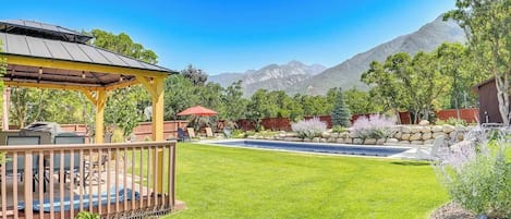 A backyard with a wooden deck, a barbecue area, a swimming pool, and a mountain view.