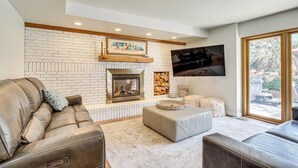 A living room with a fireplace, a sofa, ottomans, and a TV mounted on the wall. - Sandy East Bench • Pool • Mountain Views • Hot Tub (Sandy)