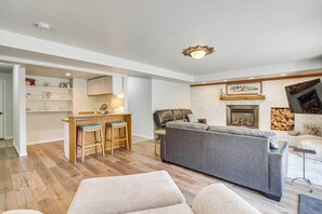 A modern living room with a grey sofa, wooden bar stools, a fireplace, and a kitchen area with white cabinets and a brick wall. - Sandy East Bench • Pool • Mountain Views • Hot Tub (Sandy)