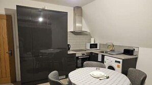 A compact kitchen with a refrigerator, microwave, and sink. There is a round table with chairs and a small countertop with a kettle and a toaster. - Affordable 2 bedroom flat in Bedford (Bedford)