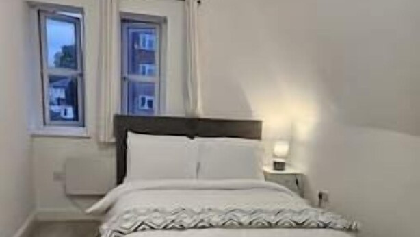 A bedroom with a bed, a nightstand, a window with curtains, and a patterned rug. - Affordable 2 bedroom flat in Bedford (Bedford)
