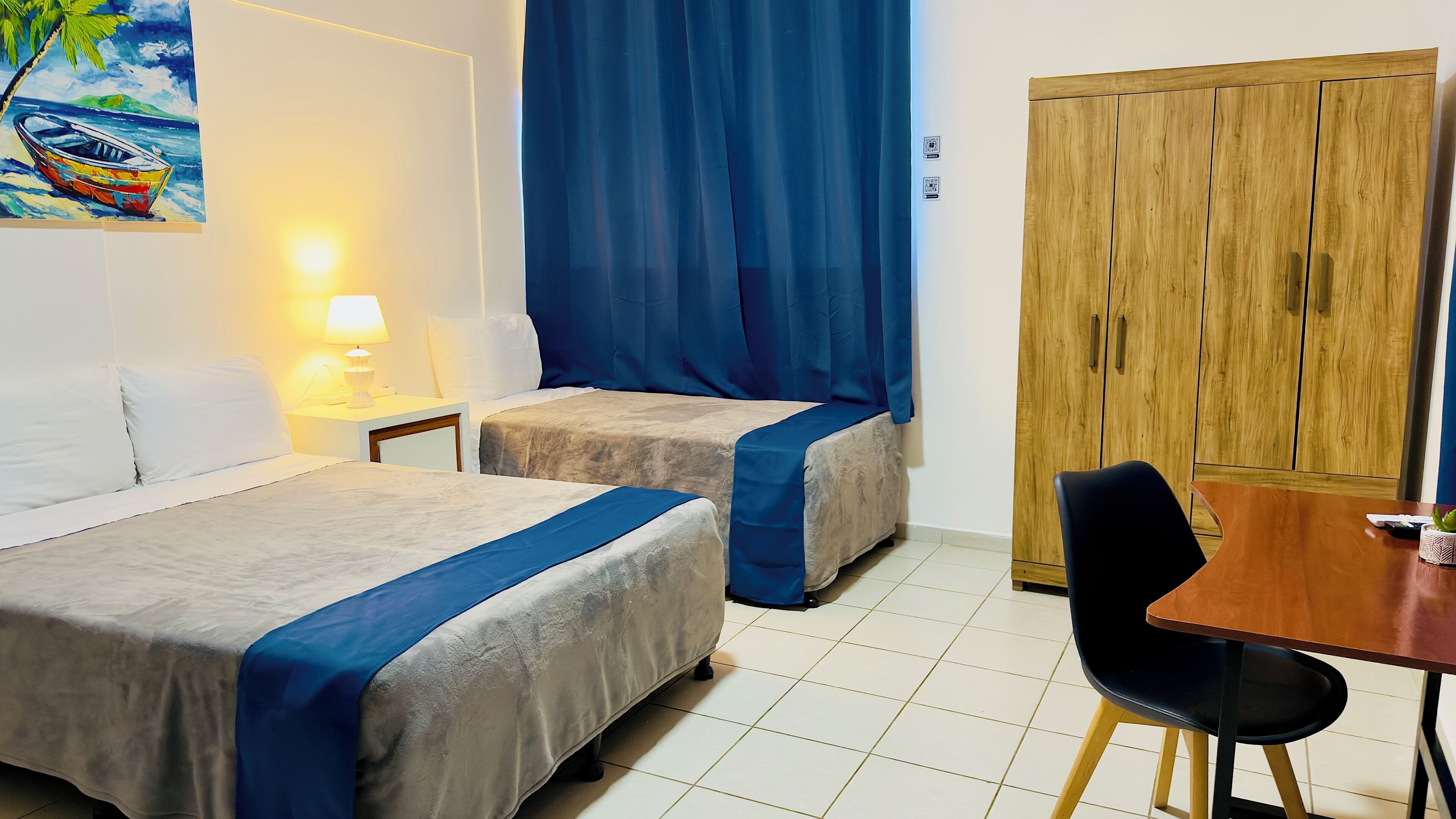 A hotel room with two beds, a desk, a chair, a wardrobe, and a painting on the wall.