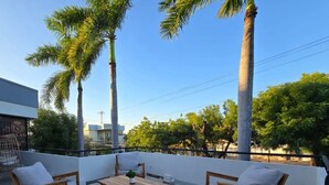 Una terraza en la azotea con mobiliario de madera, palmeras y vistas a la ciudad.