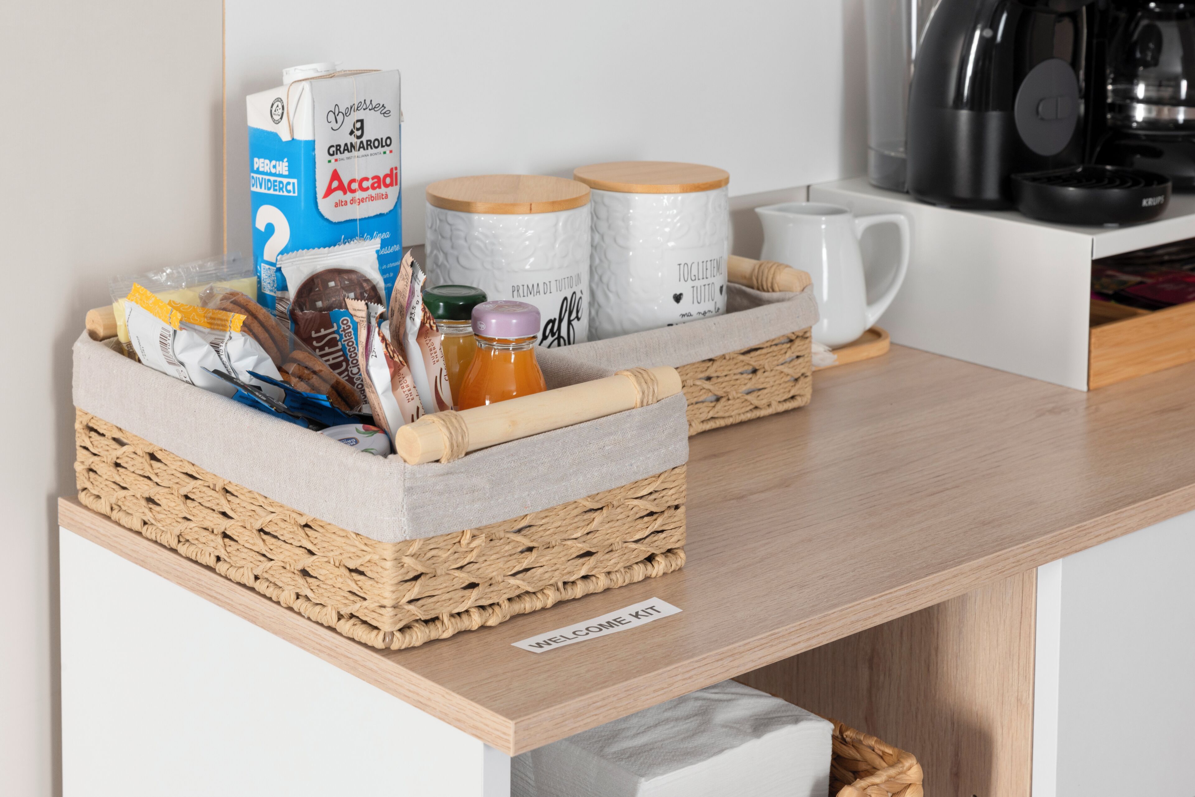 Un kit de bienvenue comprenant un carton de lait, des pots de confiture, une tasse et un panier de collations, le tout posé sur une table en bois.