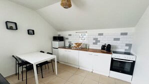Une cuisine compacte avec des meubles blancs, un plan de travail blanc, un four à micro-ondes, une cuisinière et un évier. On y trouve une table à manger avec des chaises et un luminaire suspendu.