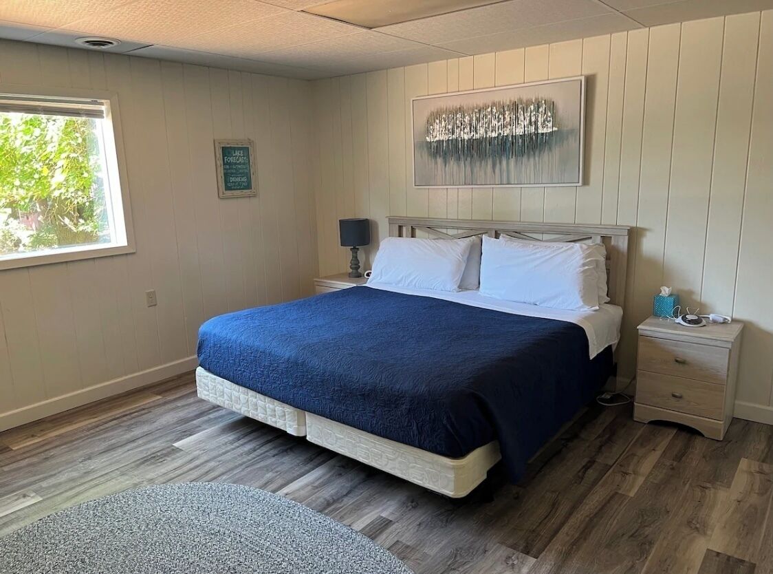 A bedroom with a bed, a nightstand, a lamp, a framed picture, and a window with a view of greenery.