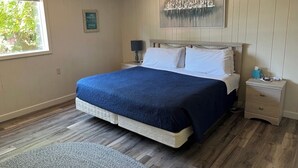 A bedroom with a bed, a nightstand, a lamp, a framed picture, and a window with a view of greenery.
