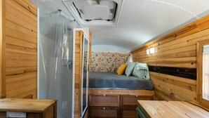 A compact RV interior with a wooden interior, a bed with a floral bedspread, and a small kitchenette area. - The Pinnacle Mountain Bus Stop ~ Fire pit, Frisbee Golf, Baggo, Hammock (Little Rock)