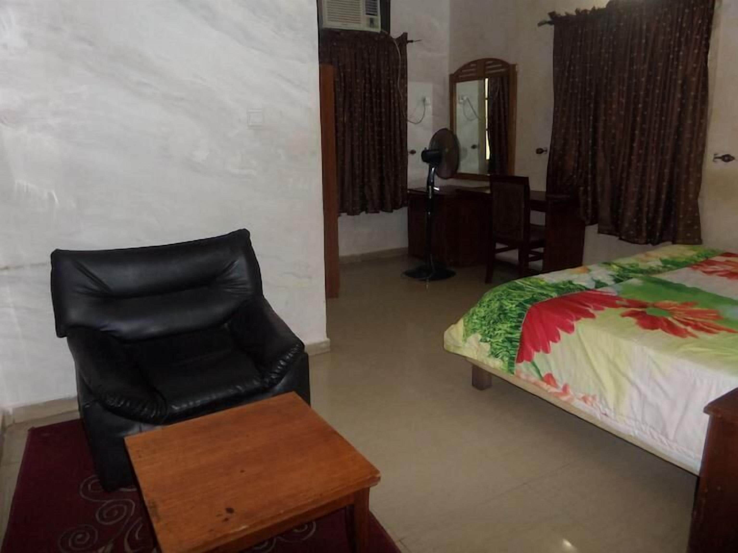 A room with a bed, a chair, a small wooden table, a desk with a mirror, and a window with curtains.