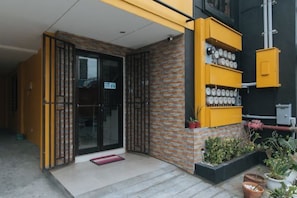 A building entrance with a glass door, a yellow wall with shelves, and a small garden area. - 4720 Silverio Suites Paranaque City (Parañaque)
