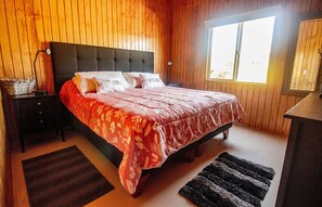 A bedroom with a bed, wooden walls, a nightstand, a lamp, and a window. - Oceana Suites Curiñanco (Valdivia)