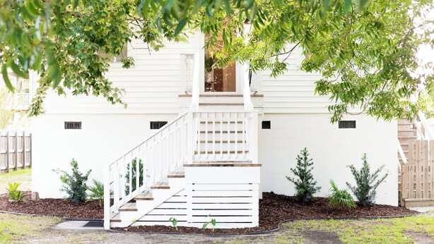 A white building with a staircase leading to a second-floor balcony, surrounded by small trees and shrubs.