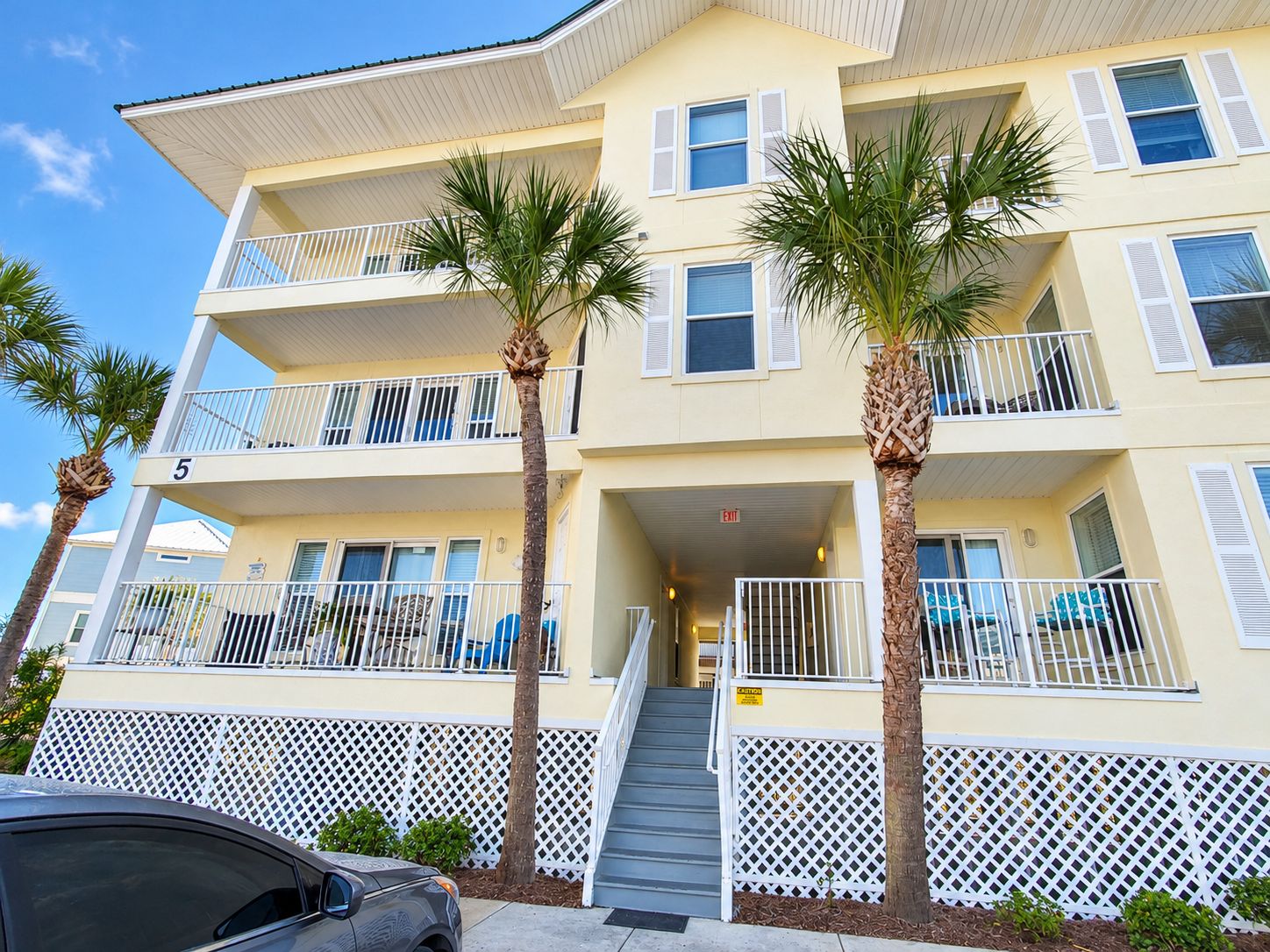 A multi-story hotel with balconies, palm trees, and a staircase leading to the entrance.