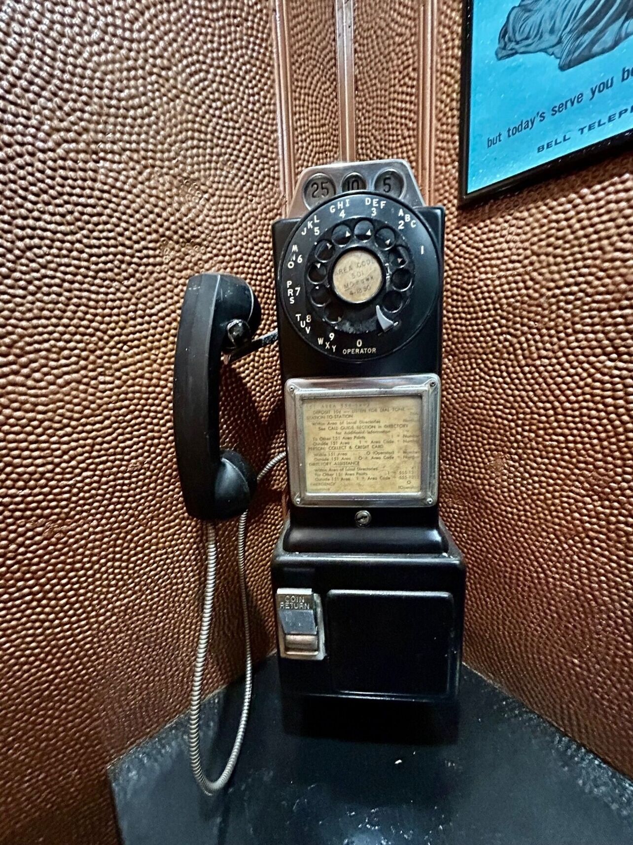 A vintage rotary dial telephone with a coin return slot, mounted on a textured brown wall.