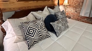 A bed with a white quilt and patterned pillows, a wooden headboard, a painting above the bed, and a brick wall. - Room 4 (sleeps 2) at Chipley's Squareview Inn (Carthage)