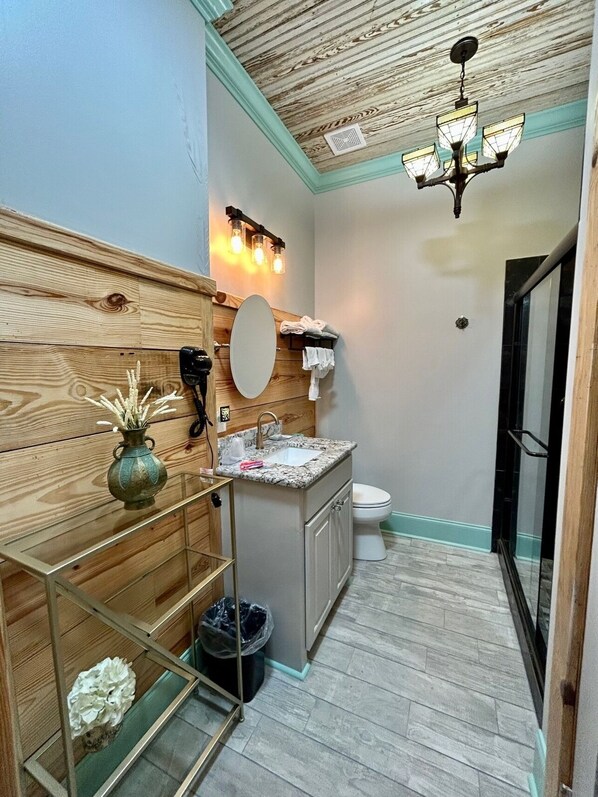A compact bathroom with a wooden vanity, a sink with a marble countertop, a mirror, a toilet, and a shower area. - Room 5 (sleeps 2) at Chipley's Squareview Inn (Carthage)