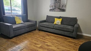 A living room with two grey sofas, one with a yellow pillow and the other with a white flower pillow, and a wooden floor. - Welcome to our inviting 2-bedroom home, perfect for accommodating up to 6 guests. Located just minutes from the Airport and McKinney Falls, this triplex-style home offers a comfortable stay with a private entrance. (Austin)