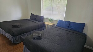 Two single beds with dark bedding, a small bedside table with a plant, and a window with blinds. - Welcome to our inviting 2-bedroom home, perfect for accommodating up to 6 guests. Located just minutes from the Airport and McKinney Falls, this triplex-style home offers a comfortable stay with a private entrance. (Austin)