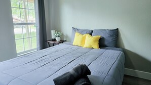 Un letto rifatto con un piumone grigio e due cuscini, uno giallo e uno grigio, in una stanza con una finestra e un comodino con una pianta.
