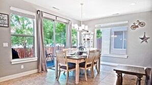 Un comedor con mesa y sillas, ventanas grandes con cortinas, una araña de cristal y vistas a zonas verdes al exterior.