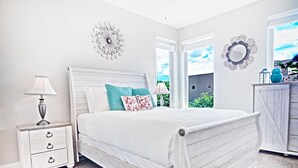Un dormitorio con una cama blanca, una mesita de noche con lámpara, un aparador y un adorno en la pared.