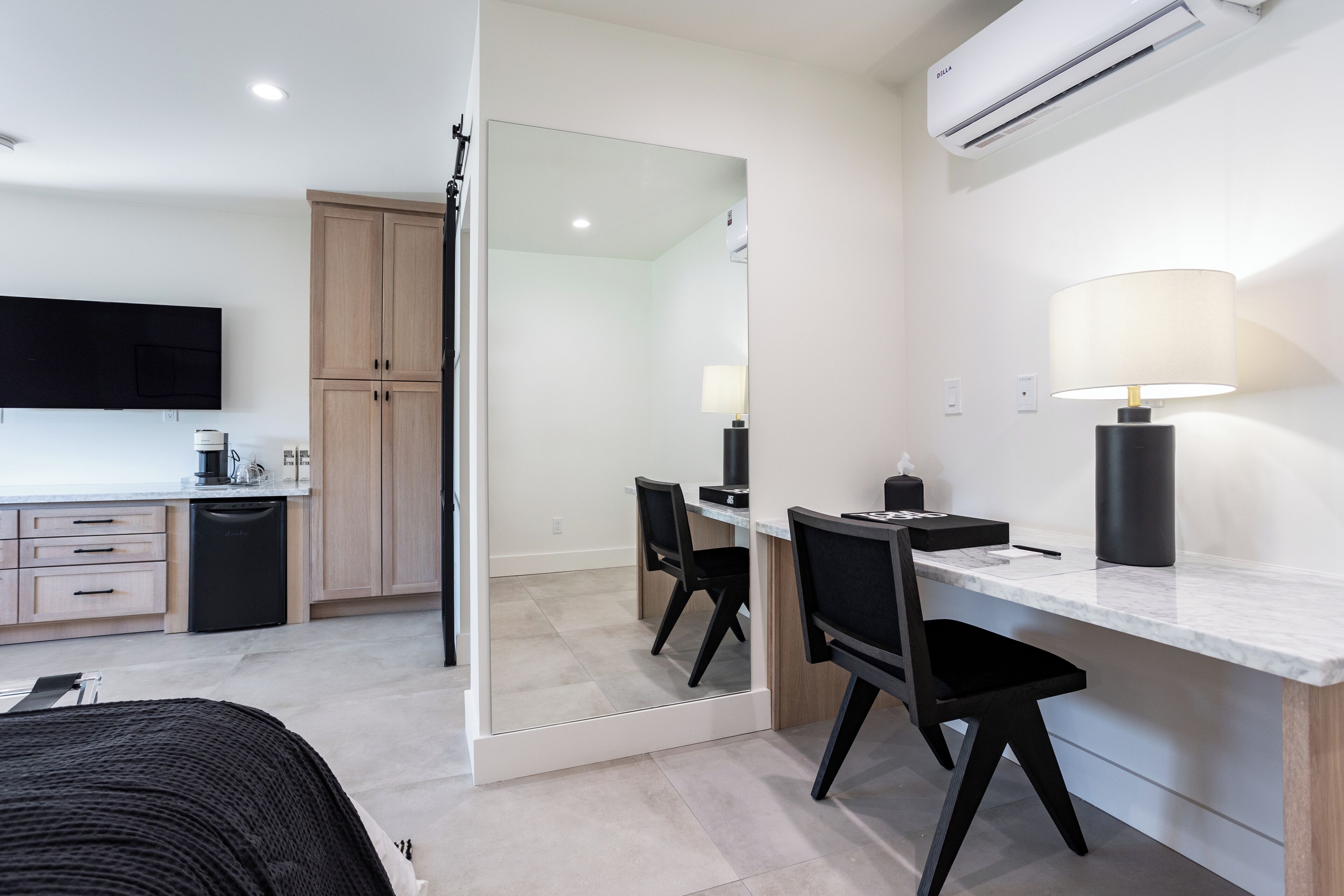 A modern hotel room with a large mirror, a desk with a lamp, a bed, and a kitchenette.