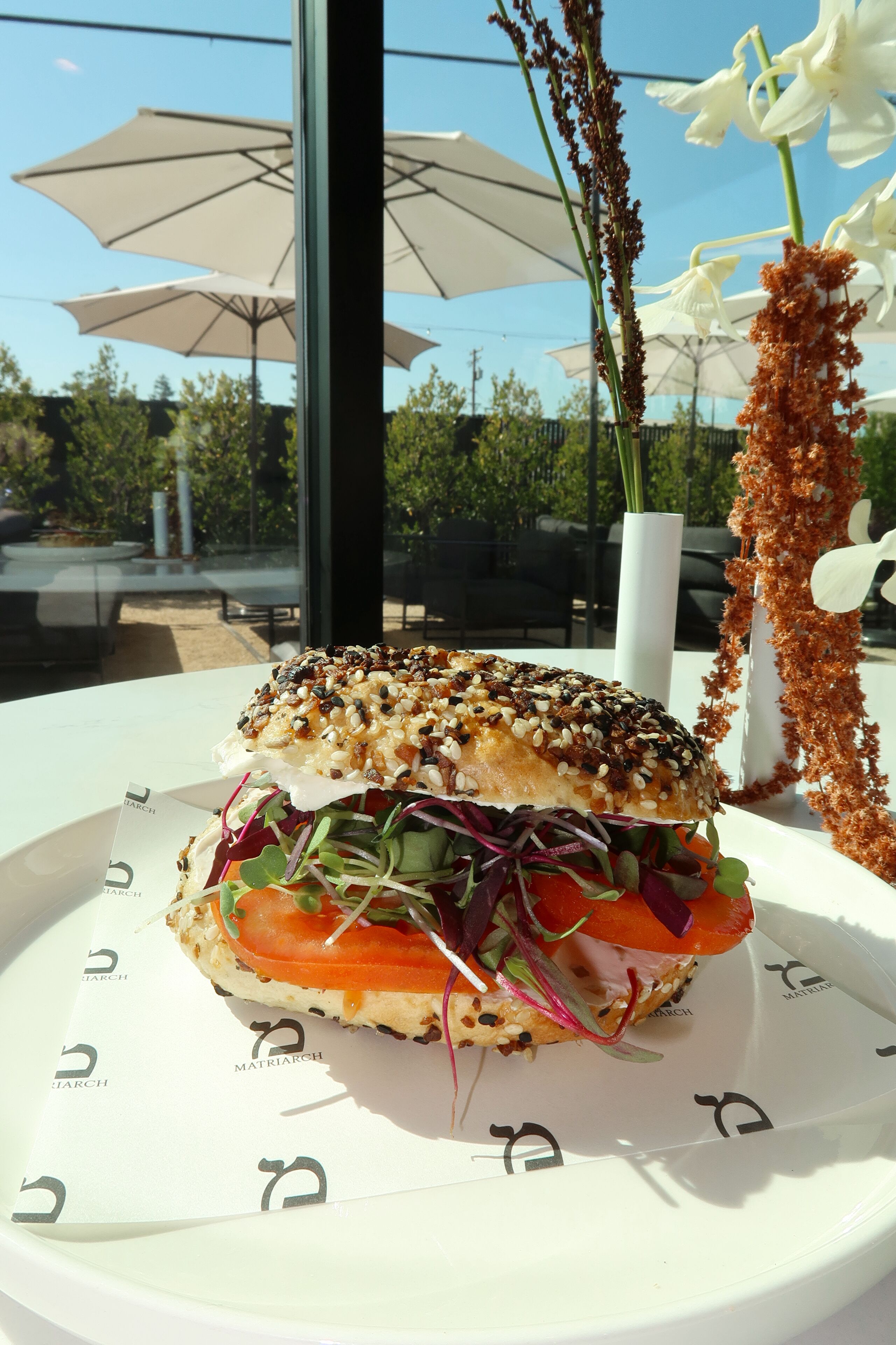 A sandwich on a white plate with a logo, a tall vase with flowers, and outdoor umbrellas in the background.