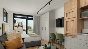 A modern living room with a sofa, a coffee table with books, a TV, and a view of the city through large windows. - The Sage l Designer Studio l Balcony Views (Cape Town)