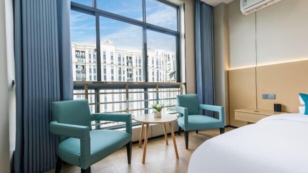 Ett hotellrum med ett stort fönster, två stolar, ett litet bord, en säng och en luftkonditioneringsanläggning.