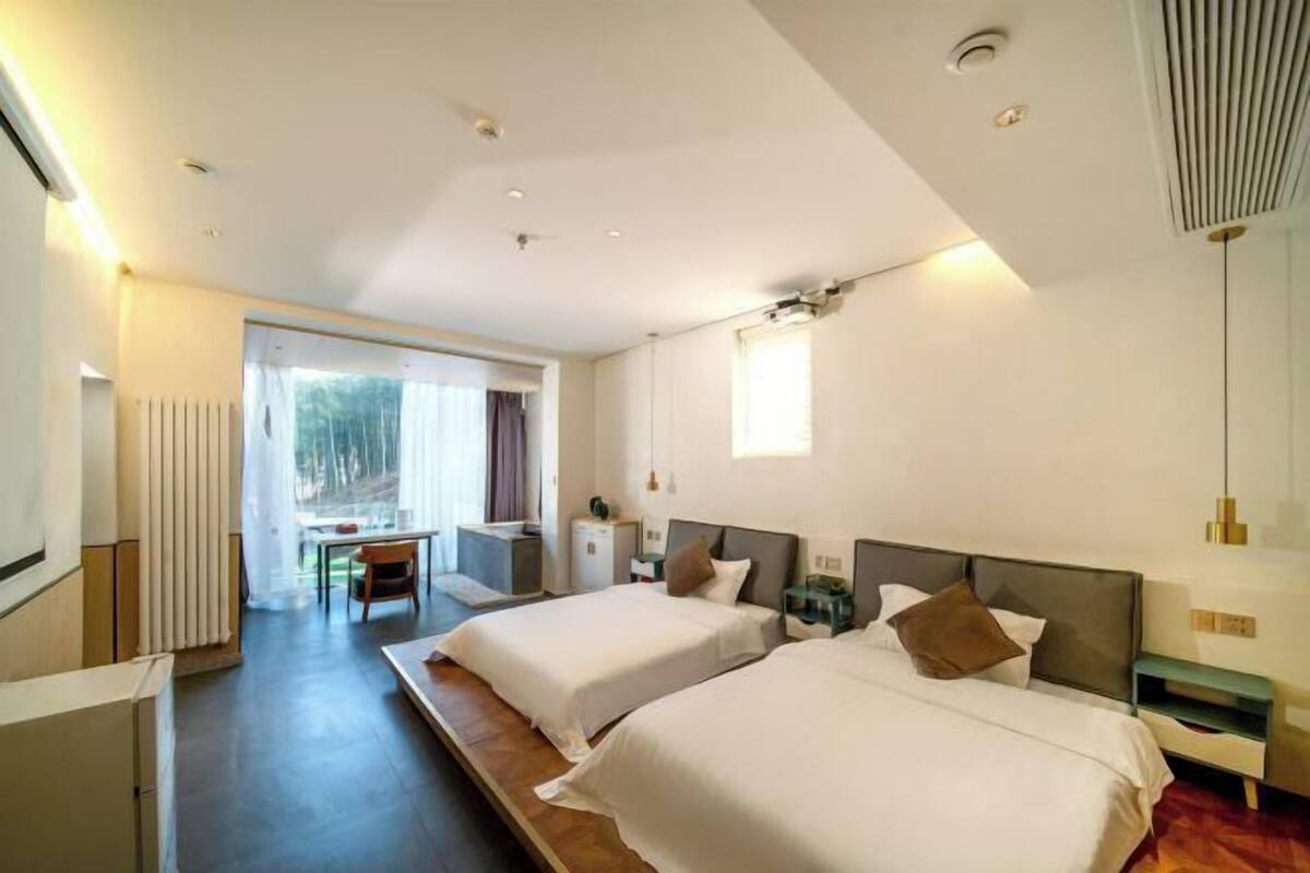 A modern hotel room with two beds, a small table, and a view of the outdoors.