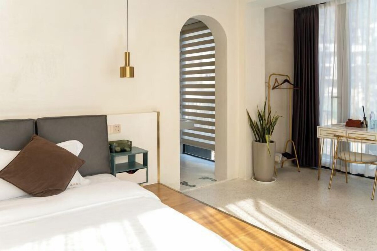 A modern hotel room with a bed, bedside table, lamp, desk, chair, and a potted plant.
