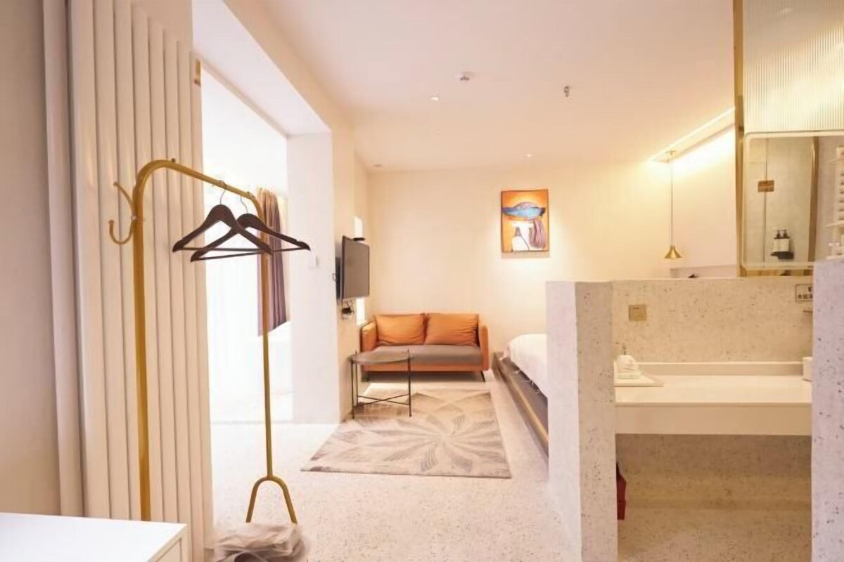 A hotel room with a bed, a sofa, a small table, a coat rack, and a bathroom with a sink and mirror.