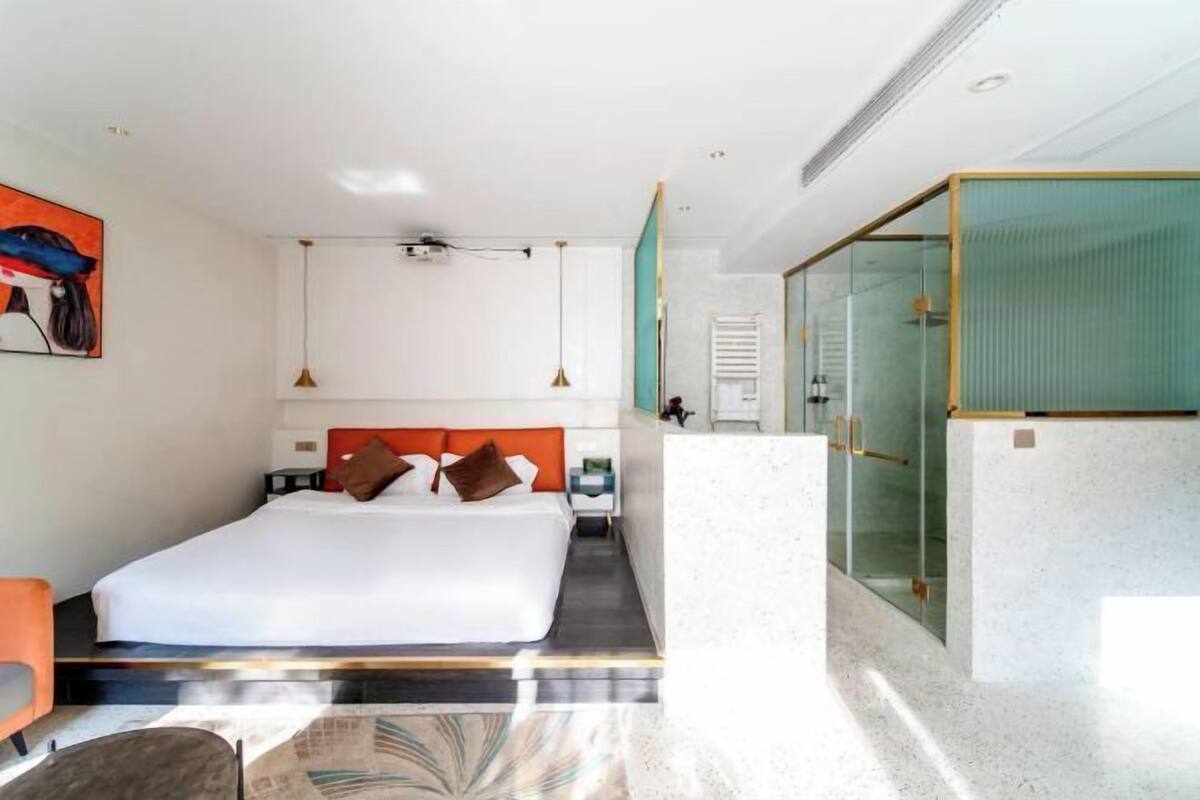 A modern hotel room with a large bed, a desk, a chair, a glass partition, and a painting on the wall.