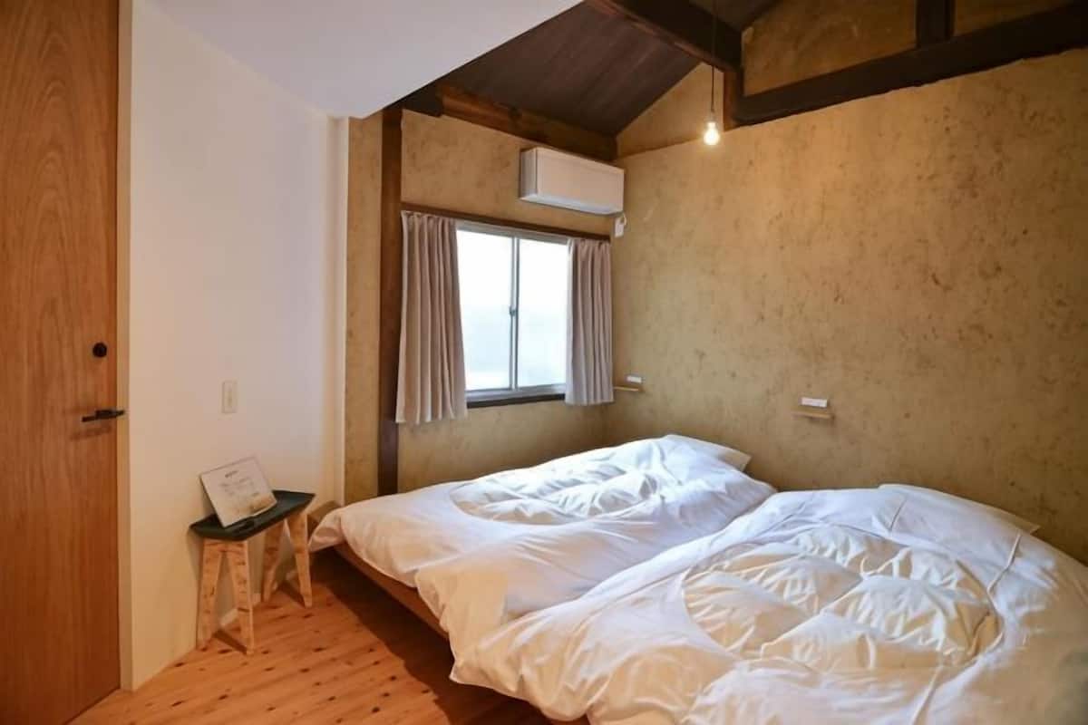 A room with two beds, a wooden floor, a window with curtains, and an air conditioning unit.
