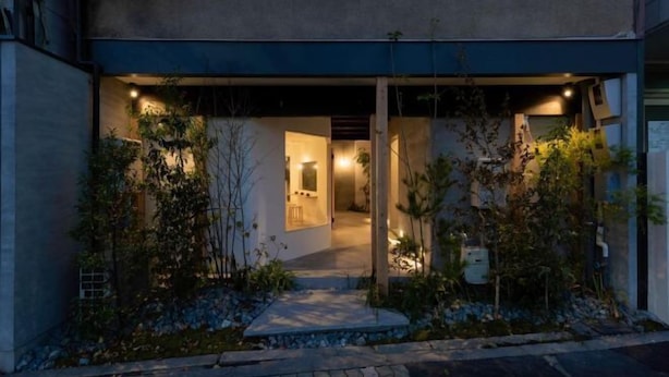 Un bâtiment moderne doté d’un hall d’entrée bien éclairé, d’un chemin menant à la porte et d’un petit jardin agrémenté de plantes et d’un élément décoratif aquatique.