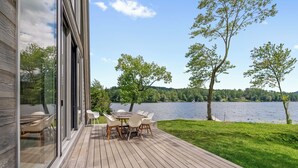 Una terraza con mesa y sillas con vista a un lago, con un área verde y árboles al fondo.