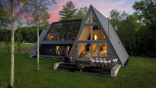 Una casa moderna de forma triangular con un amplio ventanal, un comedor con sillas blancas y una escalera que lleva a la entrada.