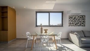 Una sala moderna con una mesa de comedor de vidrio, sillas blancas, un sofá gris y un ventanal con vista a la ciudad.