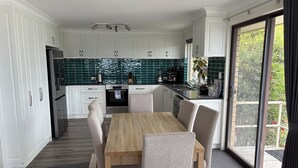 A modern kitchen with white cabinets, a wooden dining table, and a view of greenery outside. - Torndirrup Seascape - Albany (Albany)