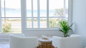 Ein Zimmer mit großem Fenster mit Blick auf einen Strand, zwei weiße Stühle, ein kleiner runder Tisch und ein Topfpflanze.