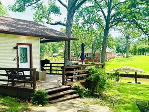 Una terraza de madera con mobiliario exterior y un porche cubierto, con vista a un paisaje verde con árboles.