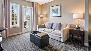 A hotel room with a beige sofa, two black ottomans, a small table, a lamp, and a framed picture on the wall.