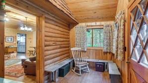 Ein Interieur einer Blockhütte mit Schaukelstuhl, Holzbänken, einem Esstisch und Blick ins Freie durch ein Fenster mit Vorhängen.