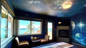 A bedroom with a bed, a window with a view, a chair, and a wall with a space-themed mural. - Shangrila  Oceanfront Retreat | 4 Ensuite Suites & 360° Views (Pender Island)
