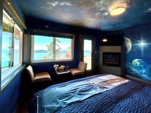 Un dormitorio con cama, ventana con vistas, una silla y un mural con temática espacial en la pared.