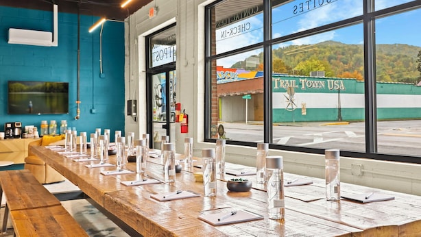 A long wooden table set with glasses and notepads, large windows with a view of a building outside, and a blue wall with a mounted TV. - The Speckled Trout Outpost (Damascus)