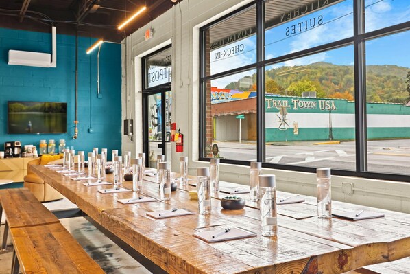 A long wooden table set with glasses and notepads, large windows with a view of a building outside, and a blue wall with a mounted TV. - The Speckled Trout Outpost (Damascus)