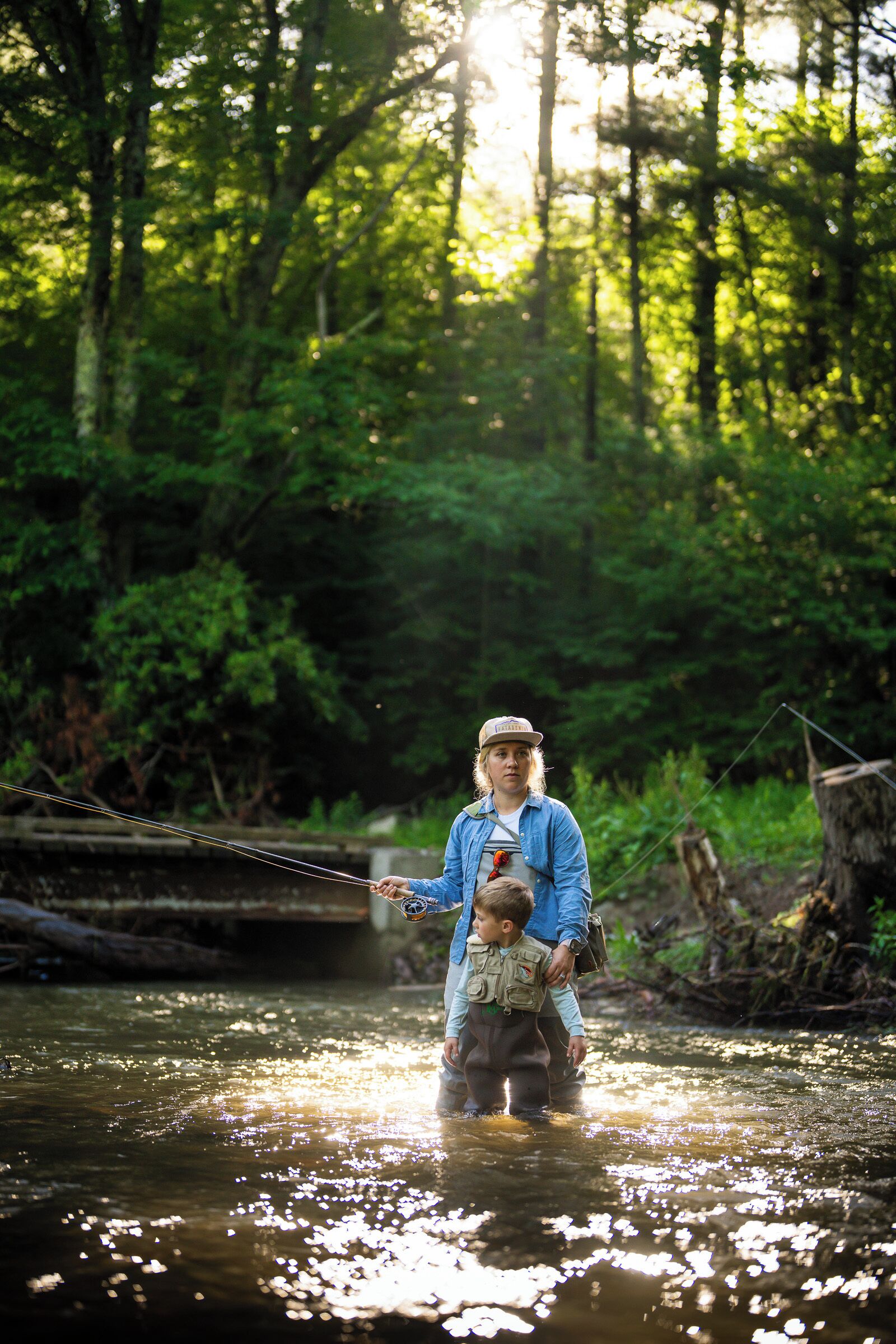 A person standing in a forested area with a river, holding a fishing rod.