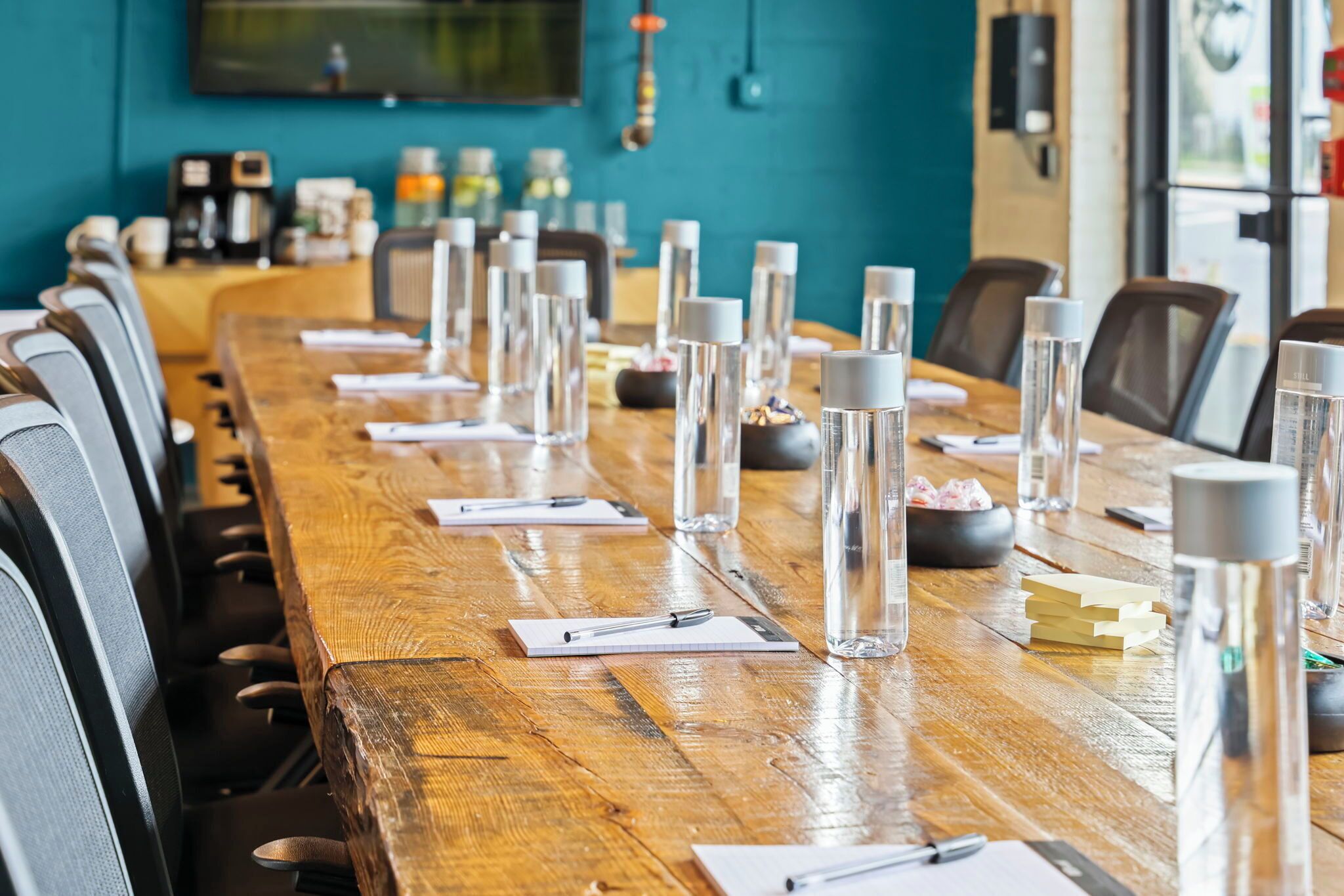 Un salón de conferencias con una larga mesa de madera, sillas, botellas de agua, cuadernos, bolígrafos y una máquina de café.